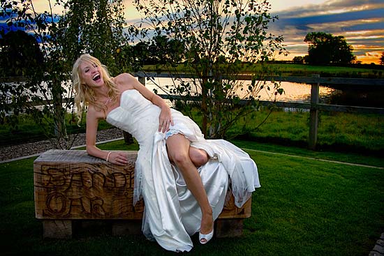 Bride sitting on wooden seat with sunset backdrop at Sandhole Farm, Cheshire.